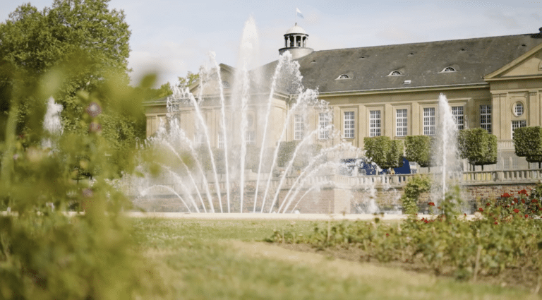 Brunnen mit Wasserfontänen vor einem historischen Gebäude in Bad Kissingen, umgeben von grünen Pflanzen und Blumen.